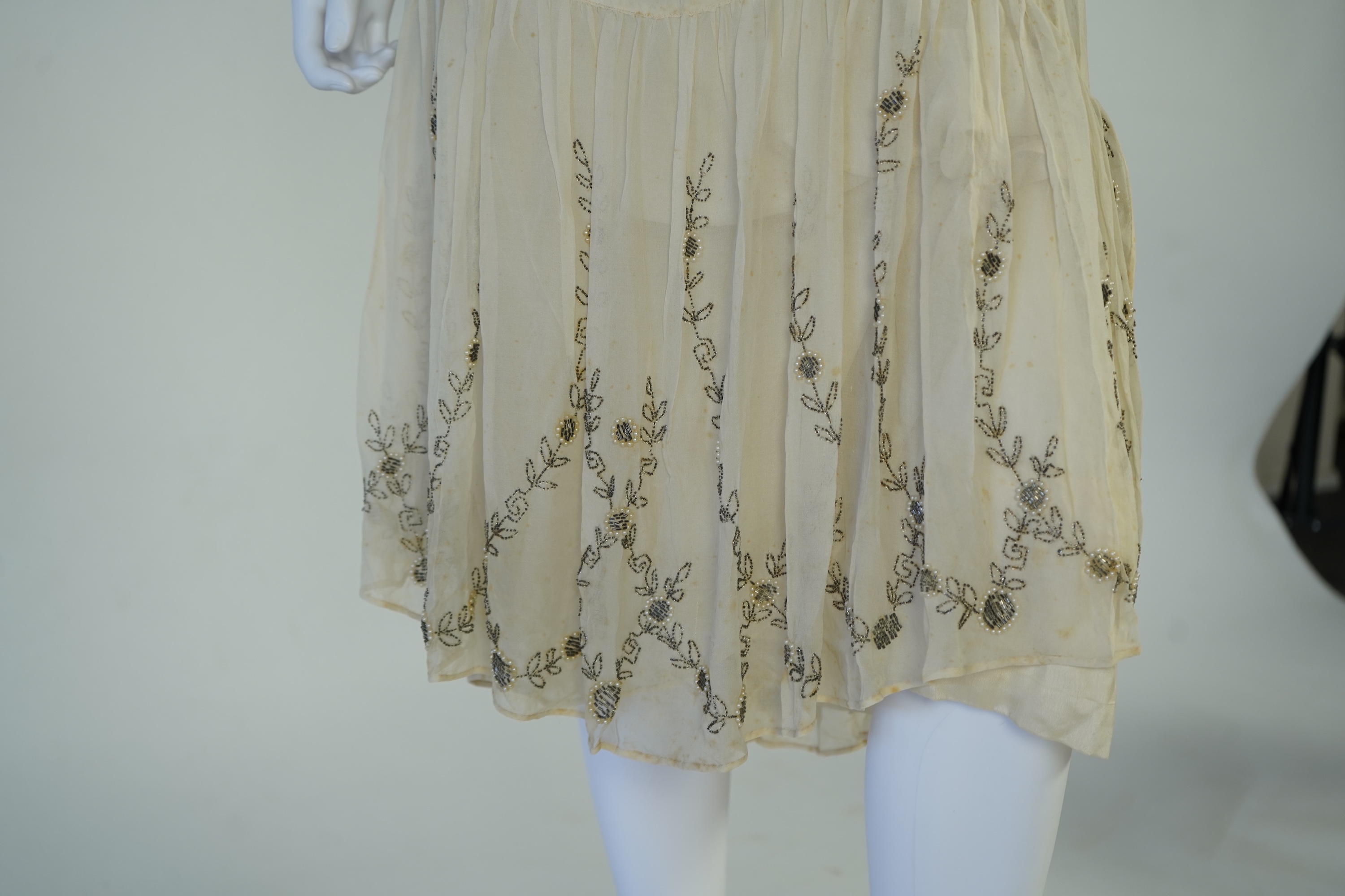 A 1920's cream silk georgette three quarter length, silk lined ‘flapper dress’. Designed with beaded and pearl floral embroidery, a stylish long beaded keyhole slit at the back, a beaded front bodice and flying beaded pa